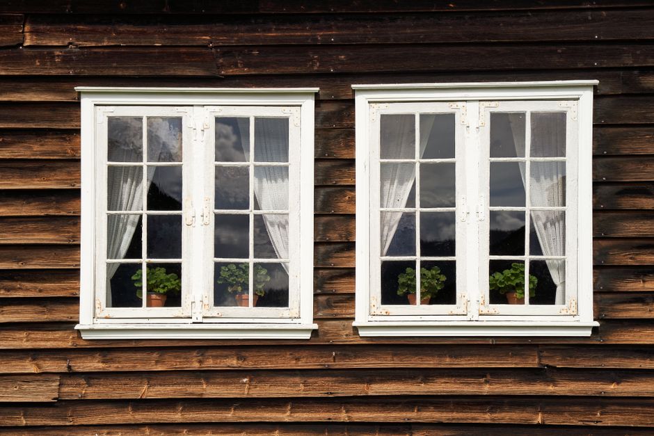 Deux fenêtres à cadre blanc sur une façade en bois, encadrées de rideaux clairs.