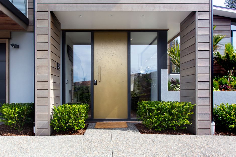 Porte d'entrée contemporaine à panneau doré et vitrages latéraux, encadrée de verdure.